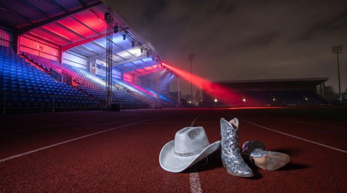 Empty floodlit stadium at night lit in red, white and blue, with a rhinestone cowboy hat and sequined boots on the track