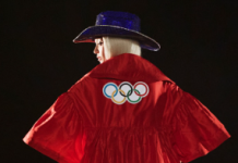 Beyoncé Shines in Surprise Olympics Tribute, Welcoming Team USA to Paris The image features Beyonce wearing a red suit with the Olympic rings emblem on the back. She is also wearing a blue, wide-brimmed hat. The scene appears to be dark, with the person illuminated against a dark background. The jacket has a loose, flowing design, and Queen B is seen from behind, slightly turned to the side.