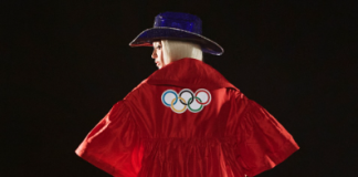 The image features Beyonce wearing a red suit with the Olympic rings emblem on the back. She is also wearing a blue, wide-brimmed hat. The scene appears to be dark, with the person illuminated against a dark background. The jacket has a loose, flowing design, and Queen B is seen from behind, slightly turned to the side.