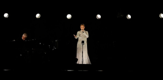 Celine Dion performing at the Paris 2024 Olympics, dressed in a white Dior gown, standing on the Eiffel Tower with a backdrop of the Olympic rings.