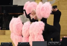 Lady Gaga’s Paris Olympics Performance: A Dazzling Showcase Lady Gaga performs at the 2024 Paris Olympics Opening Ceremony, wearing a black off-shoulder outfit and surrounded by pink feathered fans held by dancers, set on a golden staircase.