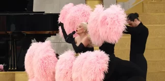 Lady Gaga performs at the 2024 Paris Olympics Opening Ceremony, wearing a black off-shoulder outfit and surrounded by pink feathered fans held by dancers, set on a golden staircase.