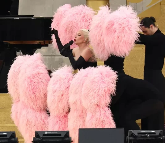 Lady Gaga’s Paris Olympics Performance: A Dazzling Showcase Lady Gaga performs at the 2024 Paris Olympics Opening Ceremony, wearing a black off-shoulder outfit and surrounded by pink feathered fans held by dancers, set on a golden staircase.