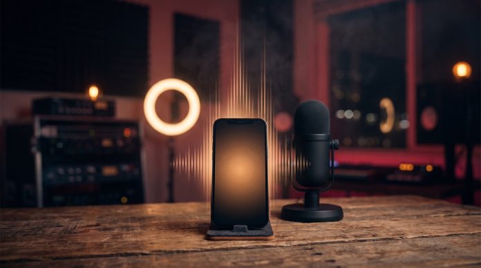 A smartphone on a home studio desk beside a microphone and ring light, with glowing sound-wave bars rising from the screen