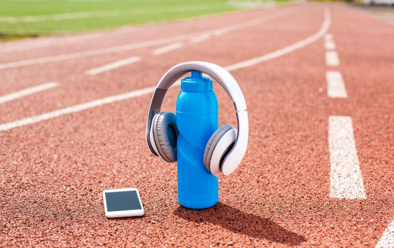 Headphones placed on a blue water bottle with a smartphone on a running track, illustrating the integration of music into athletic training routines.